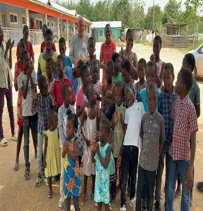 A group of children and an adult standing outdoors in front of a building at an orphanage in Ghana, smiling and clapping together.